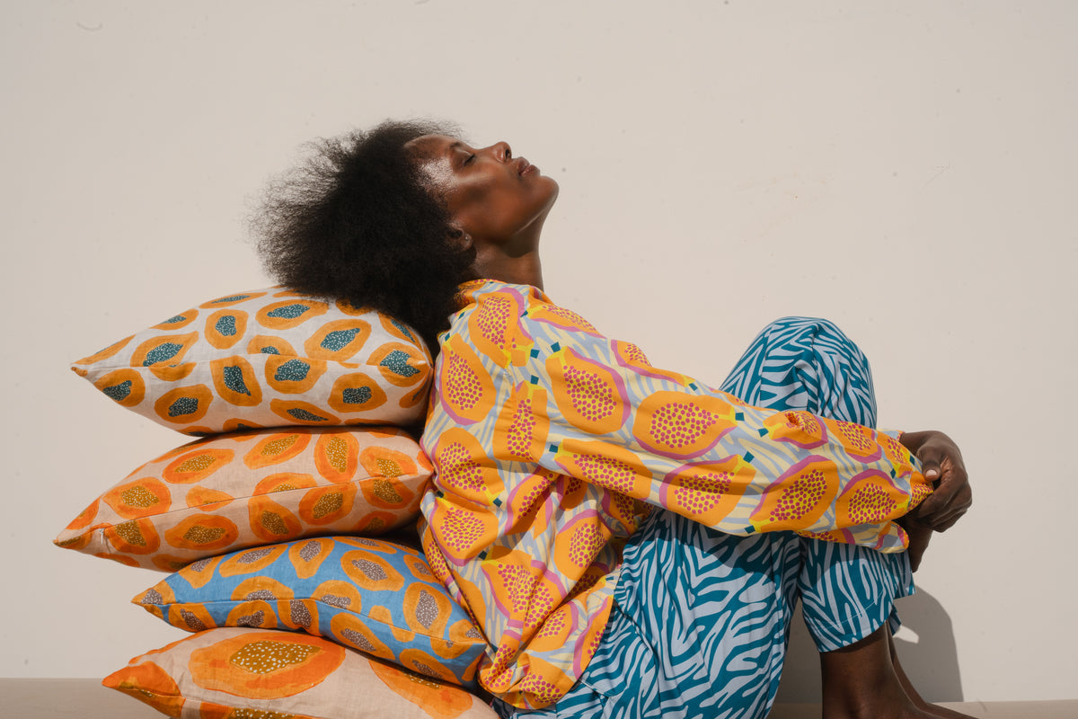 Model seated against a stack of yellow, orange and blue printed COCOLILI cushions